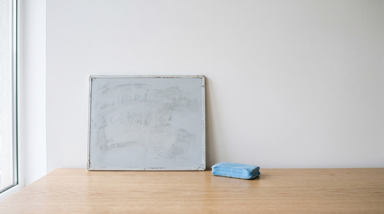 A small whiteboard wiped almost clean on a light desk, faint smudges left, a blue eraser beside it